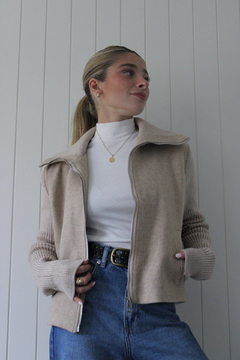 Campera Marea de color beige, con cierre frontal y mangas de punto, combinada con una camiseta blanca y jeans, exhibida en un entorno minimalista.