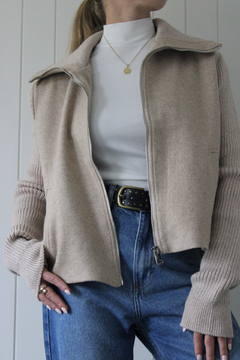 Campera Marea de color beige, con cierre frontal y mangas de punto, combinada con una camiseta blanca y jeans, exhibida en un entorno minimalista.