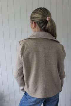 Campera Marea de color beige, con cierre frontal y mangas de punto, combinada con una camiseta blanca y jeans, exhibida en un entorno minimalista.