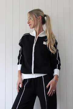 Mujer rubia con cola de caballo usando conjunto deportivo negro con detalles blancos, compuesto por campera con cierre y pantalones a juego