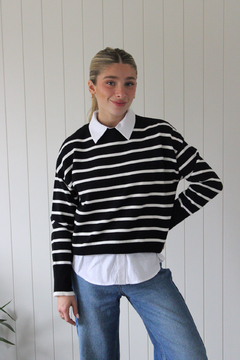 Modelo usando un sweater a rayas negras y blancas sobre una camisa blanca, posando con una mano en la cadera.