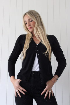 Mujer rubia con cárdigan negro Onix abotonado, camiseta blanca y pantalones negros con detalles brillantes