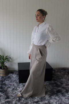 Pantalón Alhelí de color beige, combinado con una blusa blanca de mangas largas, presentado en un ambiente moderno y minimalista.