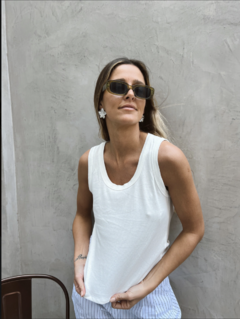 Mujer posando con musculosa blanca, gafas de sol y pendientes florales, frente a una pared gris.
