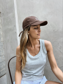 Mujer luciendo una musculosa gris con tirantes, sentada y usando una gorra marrón, con fondo de pared clara.
