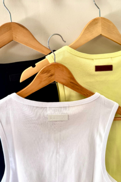 Musculosa Mirror en colores blanco, negro y amarillo, colgadas en perchas de madera.
