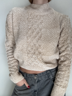 Sweater Pluma beige con detalles de trenzado, cuello alto y mangas acanaladas, combinado con pantalones oscuros.