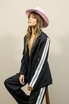 Mujer sentada con conjunto negro de pantalón y blazer con franjas blancas, sombrero rosa y accesorios dorados