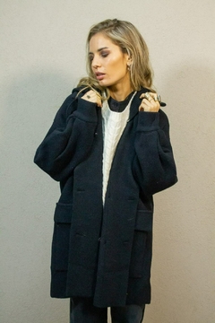 Mujer rubia usando un tapado negro oversize con solapas amplias y bolsillos grandes, combinado con un suéter tejido blanco