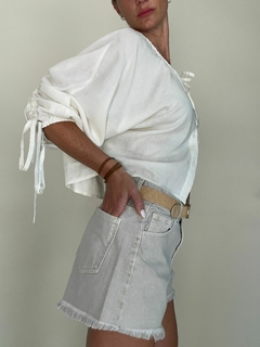 Mujer usando shorts cortos de denim claros y blusa blanca, con un cinturón beige. Fondo neutro.