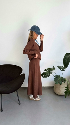 Mujer de perfil vistiendo un conjunto marrón, con una camiseta de manga larga y una falda larga, usando una gorra azul y zapatillas deportivas. Al fondo, una planta y un sillón negro.
