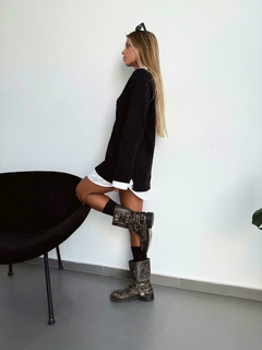 Mujer luciendo un sweater negro de la nueva colección Otoño/Invierno, con una camisa blanca debajo y botas de estilo. Fondo minimalista.