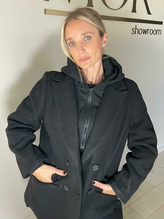Modelo usando un tapado negro Arce, posando en un showroom con paredes claras. Tapado de paño corte medio, estilo elegante-sport. Combinación de tapado con solapa y botones, campera interior con cierre conectada con la capucha (ambas desmontables). Interi