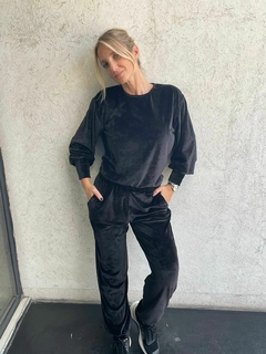 Mujer posando con conjunto deportivo negro de terciopelo, camiseta de manga larga y pantalones, con zapatillas negras y fondo de pared texturada clara