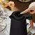 Chaleira elétrica preta com botão digital, sendo acionada por uma mão, em uma mesa com flores e itens de café.