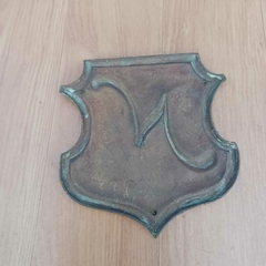 Peça decorativa em bronze com formato de escudo clássico e a letra “N” na internet