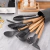 Jogo de utensílios de cozinha em silicone com 12 peças, incluindo colheres, espátulas e batedor, dispostos em um recipiente cinza ao lado de ovos e um forno.