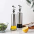 Conjunto de porta azeite e vinagre com bico dosador em aço inox, com azeite e vinagre em frascos transparentes, ao lado de uma tigela de salada e um limão cortado.