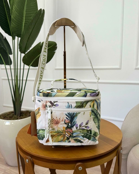 Bolsa térmica pequena com estampa de flores tropicais, usada como cooler, colocada sobre mesa de madeira ao lado de uma planta grande em ambiente interno elegante