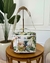 Bolsa térmica pequena com estampa de flores tropicais, usada como cooler, colocada sobre mesa de madeira ao lado de uma planta grande em ambiente interno elegante