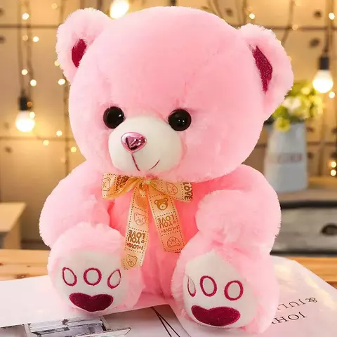 Urso de pelúcia grande rosa com laço decorativo estampado, sentado sobre superfície de madeira com fundo iluminado por luzes decorativas
