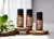 Frascos de óleo essencial da Via Aroma, incluindo Elemi, Capim Limão e Gerânio Rosa, dispostos sobre suportes de madeira com folhas verdes ao lado.