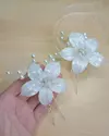 Grampo Lilly com flores brancas de resina e pérolas, segurado na mão, em fundo de mesa clara. Possui detalhes em zircônias, tamanho médio, fixação em grampo leve e grande. Produto artesanal. Ateliê Mabelly Angels Celebre.

