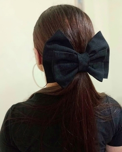 Laço Melina na cor preta, preso ao cabelo de uma mulher de cabelos longos, visto de trás. Acessório artesanal para cabelo, confeccionado em linho, com presilha de fecho regulável. Modelo de laço grande e elegante. Ateliê Mabelly Angels Celebre.