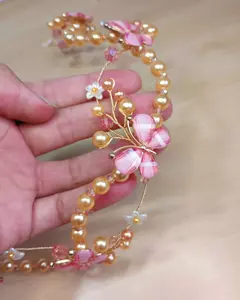 Tiara Elo Infinito, infantil e delicada. Adornada com borboletas rosa e pérolas douradas, sendo segurada por uma mão. Acessório Artesanal Flexível, regulável e com brilho suave. Ateliê Mabelly Angels Celebre.
