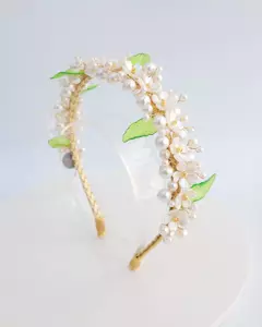 Tiara Primavera decorada com flores brancas e pérolas, com detalhes em folhas verdes, exibida em fundo neutro. Tiara infantil artesanal, para festas, casamentos. Flexível, leve e confortável. Ateliê Mabelly Angels Celebre.