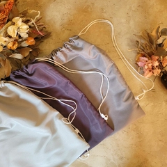 Mochila tipo saco de tecido tactel nas cores cinza claro, cinza escuro e cinza médio. Com alças que formam uma mochilas nas costas na cor  branca. Mochilas que são também sacolas.
