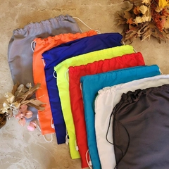 Conjunto de sacolas tipo mochilas com varias cores. Mochilas para brinde corporativos coloridas em diferentes tons, incluindo cinza, laranja, azul, verde limão, vermelho, azul claro e branco, dispostas sobre uma superfície clara.