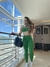 Mulher posando em um ambiente iluminado, vestindo um conjunto verde de top e leggings, segurando uma bolsa preta. Ao fundo, há uma vista de montanhas e janelas grandes. Promoção de Black Friday 