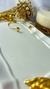 Bracelete dourado com detalhes em forma de coração, posicionado sobre uma superfície clara, próximo a um arranjo de flores secas e um copo com bebida.