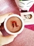 Blush compacto cor coral da marca Nina Make Up em embalagem redonda branca, com logo em relevo, sendo segurado por uma mão sobre superfície rosa brilhante, com caixa do produto ao fundo
