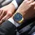 Relógio Curren masculino elegante com pulseira dourada e mostrador azul com números romanos, usado no pulso de homem vestido com camisa branca e paletó cinza