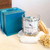 Vela aromática Cedro Real da coleção Satin Breeze, em embalagem de vidro com pedras decorativas, ao lado de uma caixa azul e pedras brancas em uma superfície clara.