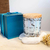 Vela aromática Cedro Real da coleção Satin Breeze, feita de cera de coco, com tampa de madeira e embalagem decorativa azul.