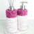 Conjunto de produtos para lavabo, incluindo um dispenser de sabonete e um difusor de ambiente, ambos com detalhes em cristais rosa e base branca texturizada.