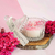 Aurora Candle in a decorative glass container, featuring a cherry blossom design and surrounded by pink flowers.