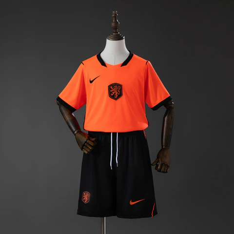 Este é o conjunto infantil titular da Holanda para a temporada 2026 da Nike, apresentando a vibrante camisa laranja com recortes pretos nas laterais e o tradicional escudo do leão da KNVB
