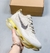 Tênis Nike Air Max Scorpion em tom claro, com detalhes em preto e sola amarela, sendo segurado por uma mão, com caixa ao fundo.