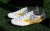 White and gold soccer cleats featuring a lion graphic design, displayed on green grass.