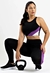 Mulher agachada usando um conjunto de moda fitness com cropped preto e calça legging preta, roxa e branca. Ao lado, um kettlebell.