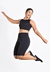 Mulher sorrindo e pulando, vestindo um conjunto fitness preto composto por cropped e bermuda.