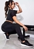 Modelo usando calça legging fitness preta com bolso em tela, agachada ao lado de um banco de treino.