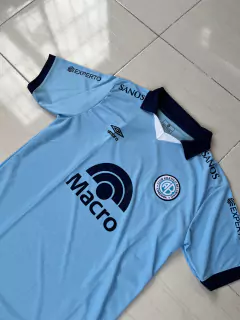 Camiseta titular de Belgrano 2024/25, color celeste, con detalles en azul marino y logotipos de sponsors.