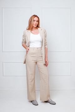 Mulher usando um blazer de linho alongado em tom bege, combinado com uma blusa branca e calças largas, em um ambiente minimalista.