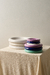 Platos decorativos Olimpia medianos apilados, en colores blanco, beige, lila, azul y verde, sobre una mesa con mantel beige