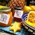 Dois potes de geleia de frutas amarelas, um com tampa dourada e outro com tampa preta, ao lado de uma tigela com frutas frescas incluindo maracujá e manga e fatias de carambola. Ao fundo, um abacaxi e uma toalha colorida.